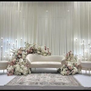 كوشة زفاف مهيبة تتوسط القاعة محاطة بكراسٍ بيضاء مُزينة بربطات زهرية، كل شيء يعكس أجواء الاحتفال والبهجة